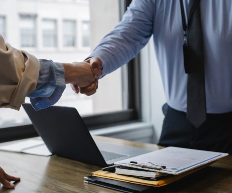 Crop unrecognizable coworkers in formal wear standing at table with laptop and documents while greeting each other before meeting